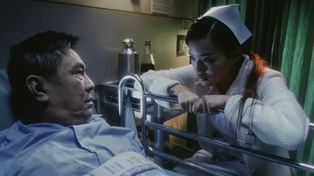 Visible Secret (2001) movie still: medium — A dimly lit hospital room features a metal bed rail and medical equipment. A nurse rests her arms on the…