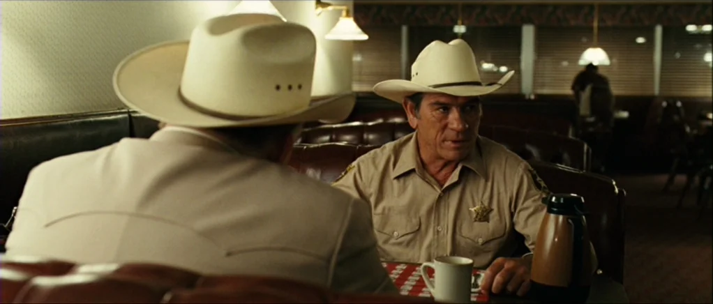No Country For Old Men movie still: medium — A dimly lit diner booth features leather seating and a small red-checkered table cloth. A sheriff in…