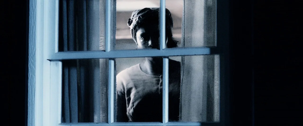 Harriet movie still: medium — A woman stands behind a wooden window frame looking out into the night. Her face is partially obscured by shadows…