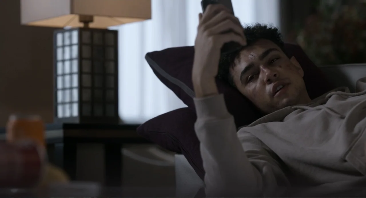 Gran Turismo (2023) movie still: medium — A contemporary living room interior with a dark sofa and a nearby table lamp. A young man lies on the sofa…