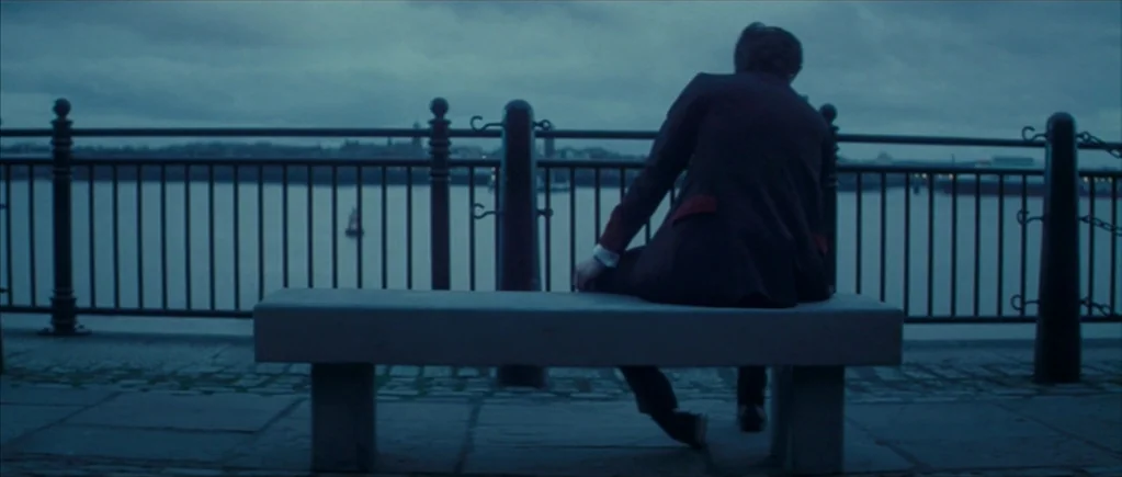 Nowhere Boy movie still: fullbody — A stone bench sits on a paved riverside promenade overlooking a wide body of water. A person in a suit sits on…