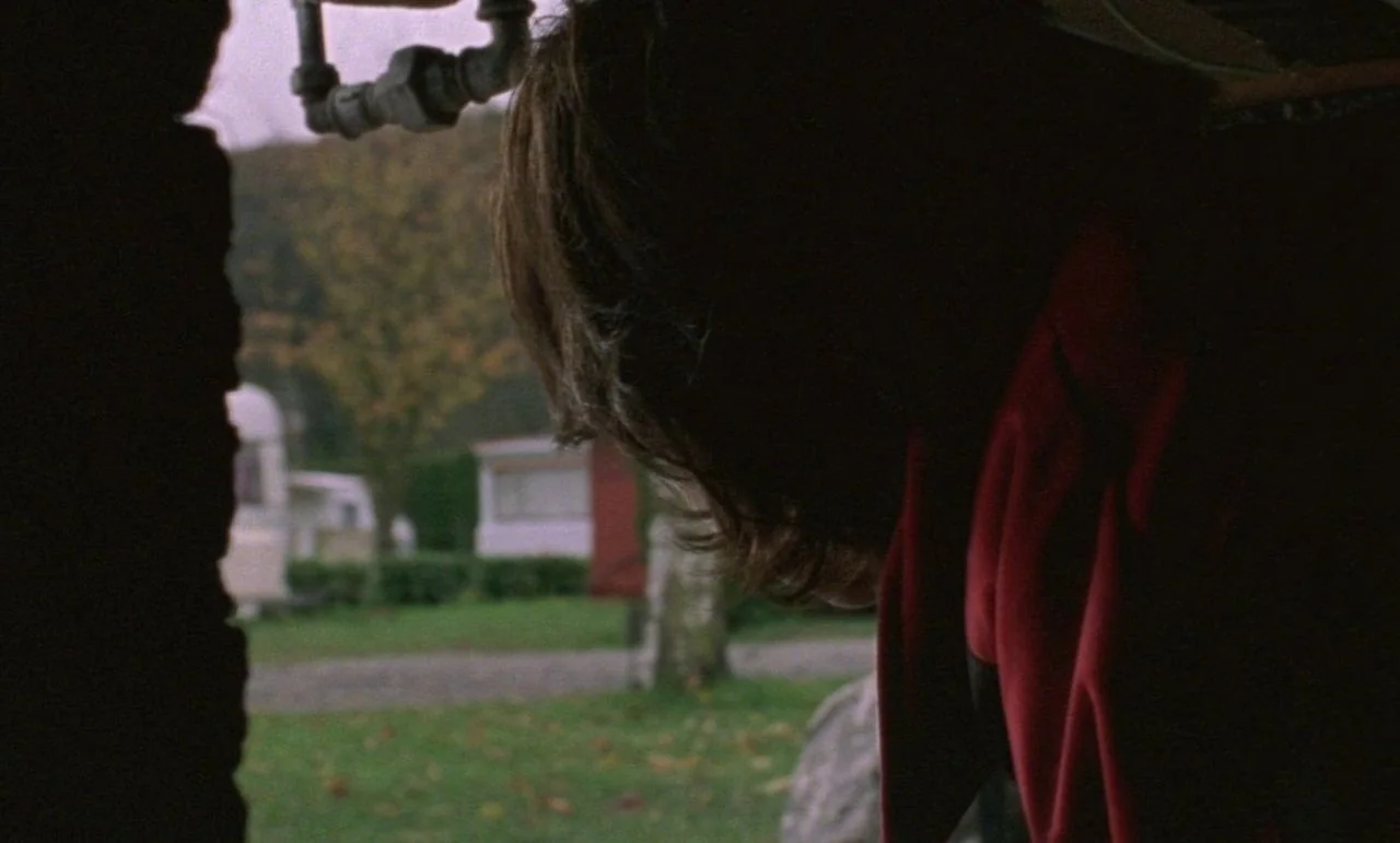 Rosetta (1999) movie still: closeup — A view from inside a structure looking out at a grassy residential trailer park. A person with messy hair in a…