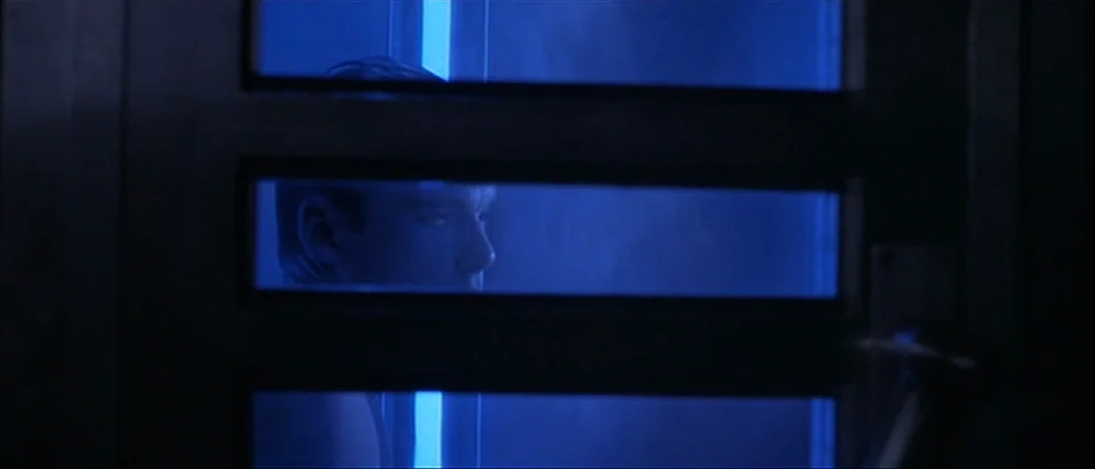 Gattaca (1997) movie still: closeup — A man is visible through the segmented frosted glass of a bathroom door. The interior is lit by a cool blue…