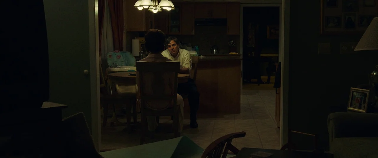 Dark Waters (2019) movie still: medium — A dimly lit room viewed from an adjacent dark space shows two people sitting. A man and a woman face each…