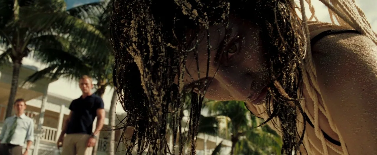 Casino Royale (2006) movie still: closeup — A woman is bound with thick white rope on a sunlit tropical beach. Two men stand in the background…