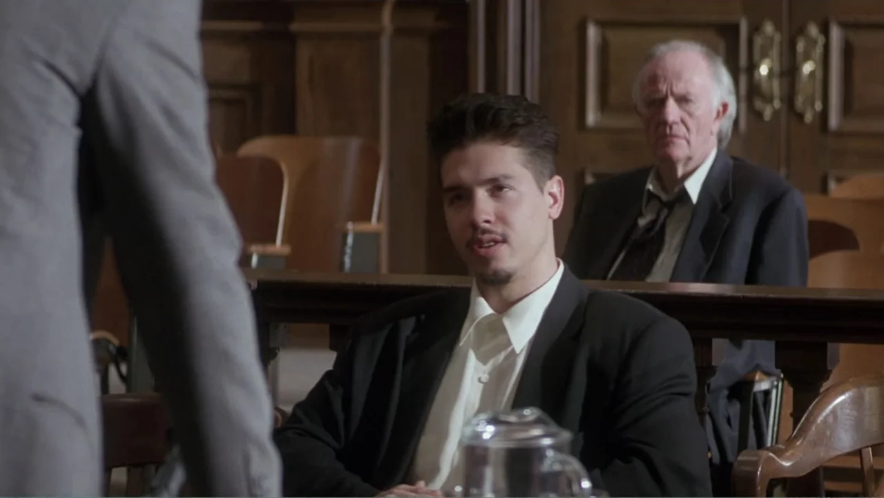 Mistrial (1996) movie still: medium — A courtroom interior features dark wood paneling and rows of empty benches. A young man sits at a table facing…