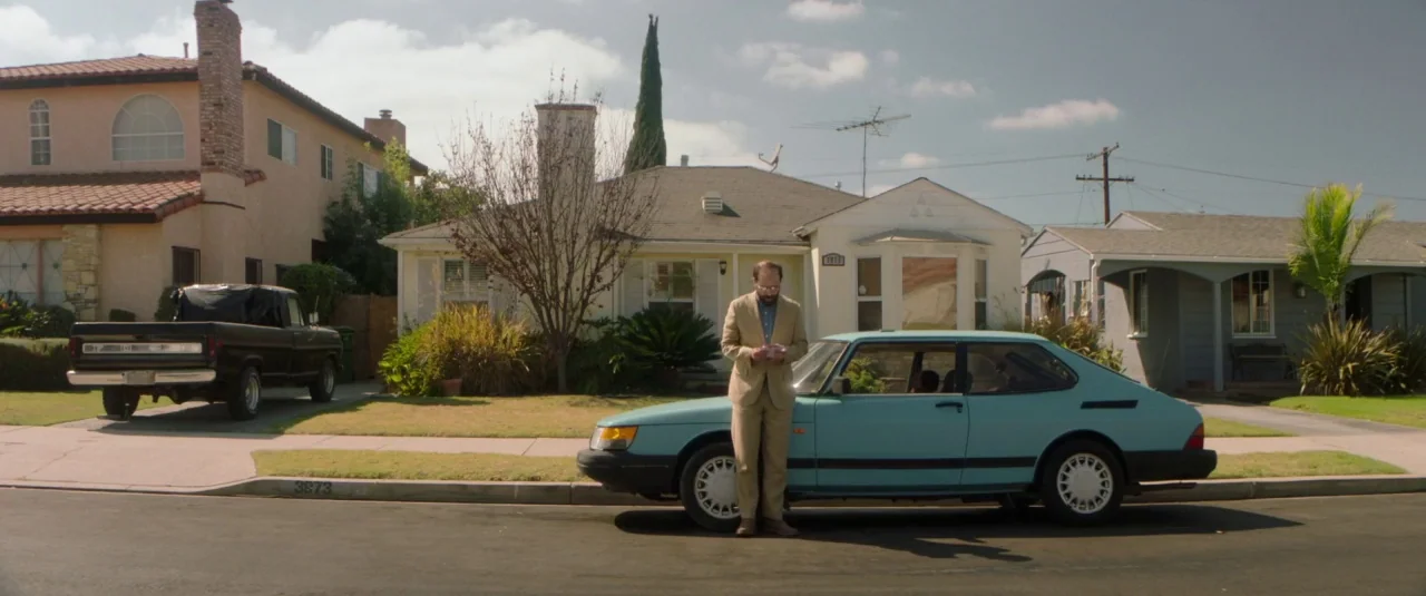 Lemon (2017) movie still: wide — A quiet suburban street with single-story houses and a parked vintage light blue car. A man in a light-colored suit…