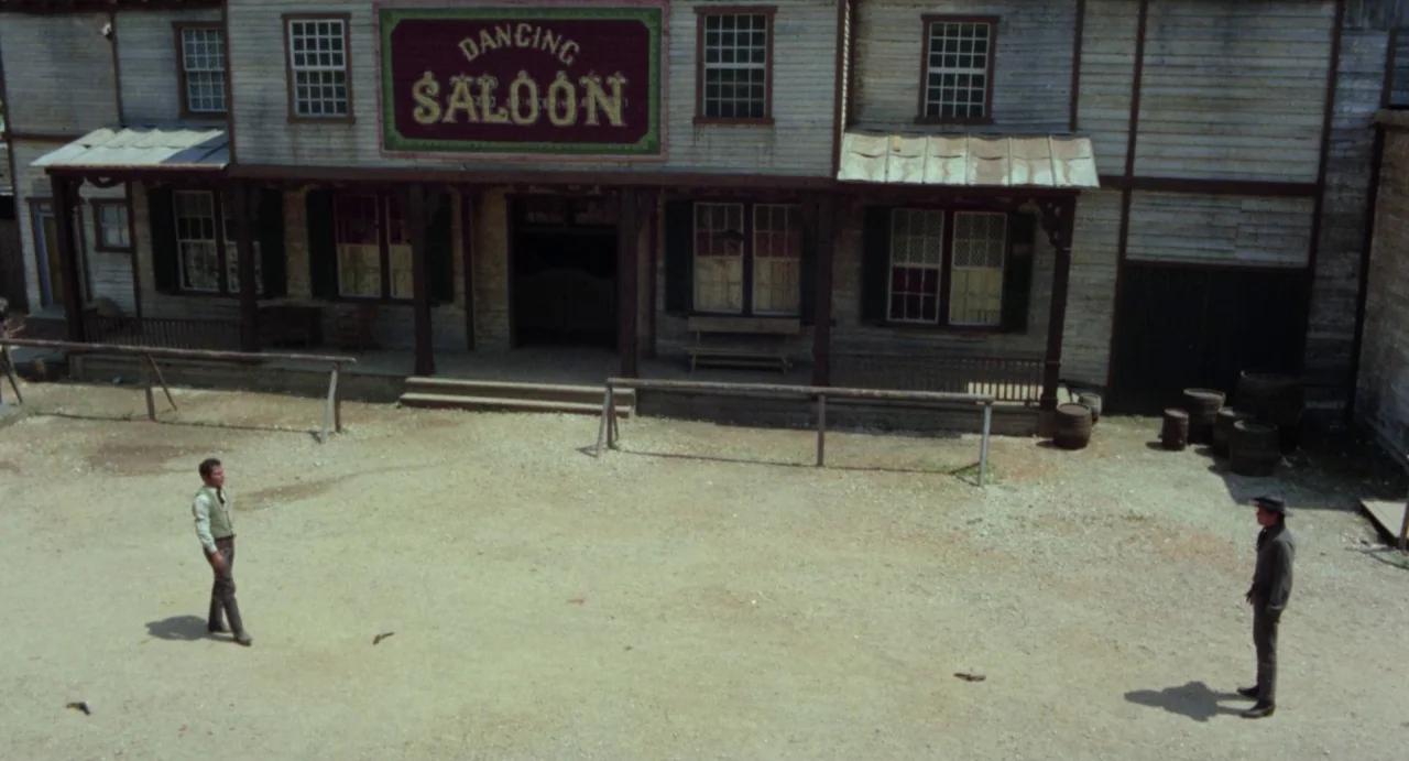 Johnny Colt (1966) movie still: wide — A dusty dirt lot sits in front of a rustic wooden building labeled Dancing Saloon. Two men stand far apart on…