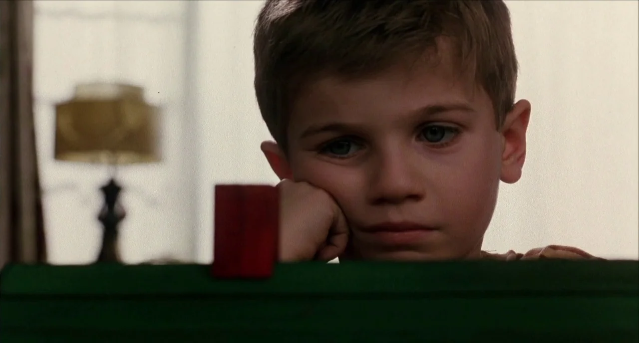 The Hours (2002) movie still: closeup — A young boy sits in a room with a green surface in the foreground. He rests his head on his hand while…