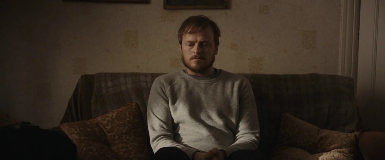 You Are Not My Mother (2021) movie still: medium — A man sits on a brown textured sofa against a wall with patterned wallpaper. The man keeps his…