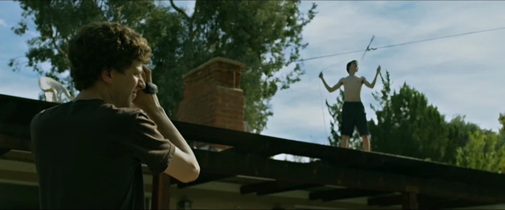 The Social Network movie still: medium — A residential rooftop with a brick chimney is set against tall green trees. One man holds a camera to his…