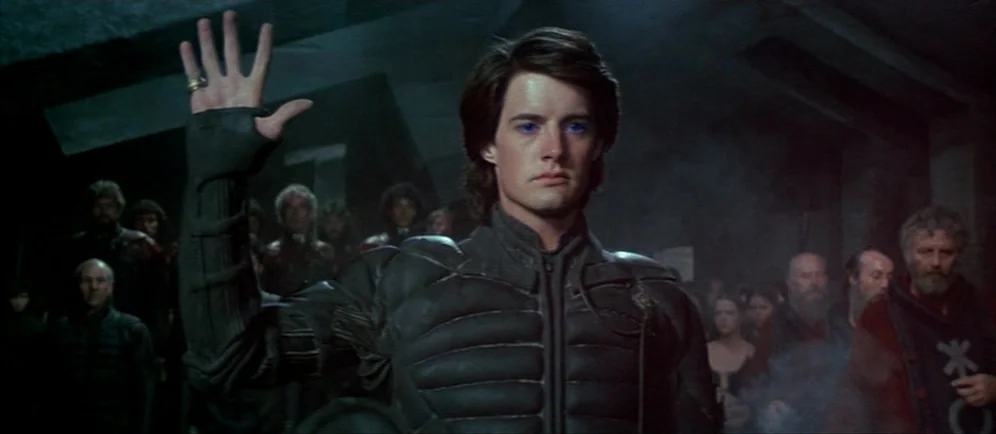 Dune (1984) movie still: medium — A dimly lit cavern filled with a crowd of people in dark, rugged clothing. A young man stands in the center…