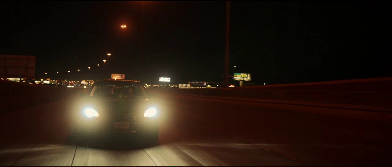 Sympathy For The Devil (2023) movie still: wide — A dark multi-lane highway at night with distant overhead lights illuminating the pavement. A…