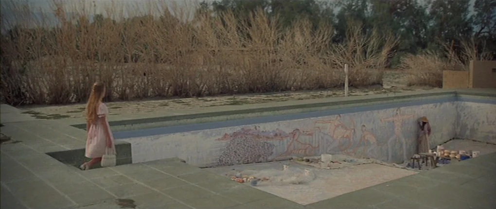 3 Women (1977) movie still: wide — A dry, empty outdoor swimming pool features murals on its interior walls. A woman in a pink dress watches as…