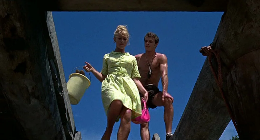 Age of Consent (1969) movie still: fullbody — A woman and a man stand on wooden beams under a clear blue sky. They appear to be walking near a…