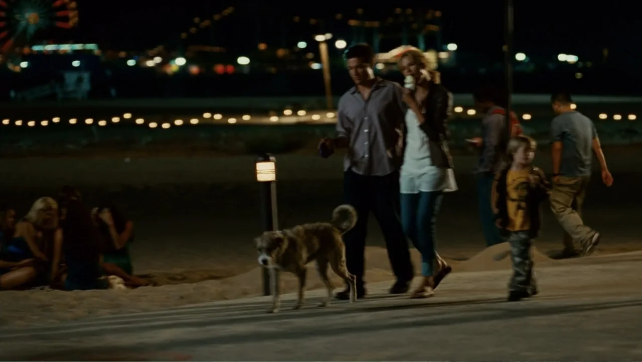 Hancock (2008) movie still: fullbody — A nighttime boardwalk area features a paved path with lamp lighting and beach beyond. A couple walks a small…