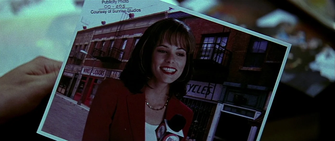 Scream 3 (2000) movie still: closeup — A close-up view shows a hand holding a glossy publicity photograph. The photograph depicts a woman in a red…
