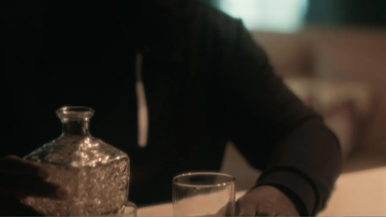SIN: Blackout (2025) movie still: medium — A dimly lit indoor space with a wooden table surface and blurred background elements. A person wearing a…
