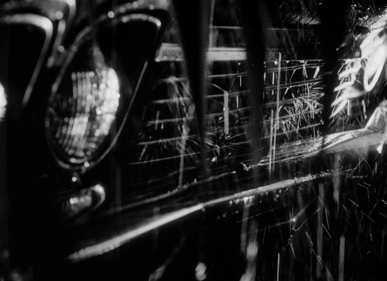 Singapore Sling (1990) movie still: closeup — A close-up view of a vintage car's chrome grille and headlight in the rain. Rain streaks blur the…