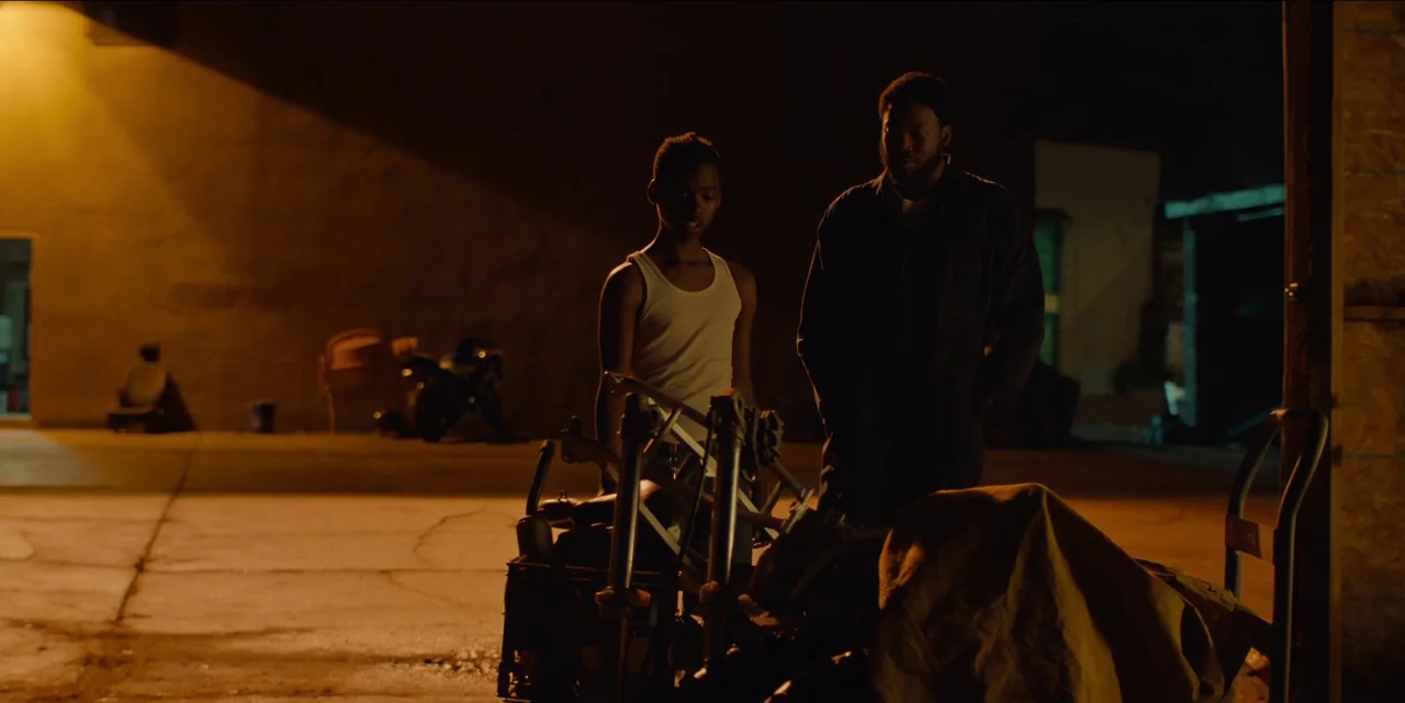 Charm City Kings (2020) movie still: fullbody — A dark outdoor concrete area illuminated by an orange street lamp at night. Two men stand beside a…