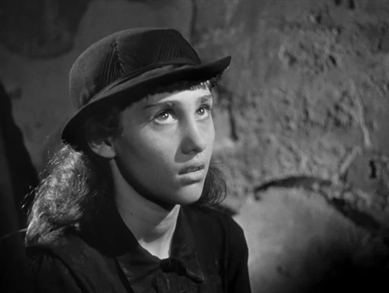 Forbidden Games (1952) movie still: closeup — A person stands against a rough, textured rock wall in a dimly lit environment. The subject looks…