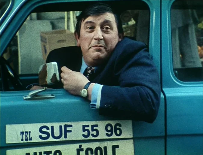 Daguerréotypes (1976) movie still: medium — A man sits in the driver seat of a blue compact car with French text. He leans his arm out of the open…