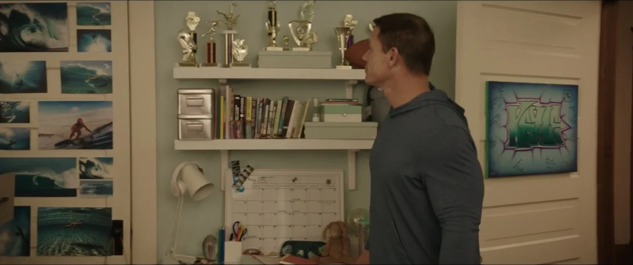 Blockers (2018) movie still: medium — A light blue bedroom wall features two white shelves filled with trophies and books. A man in a blue hooded…