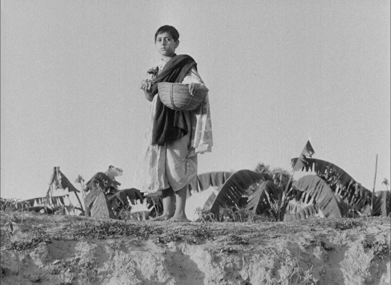 Aparajito (1956) movie still: fullbody — A rural landscape features an earthen ridge with banana trees in the distance. A young boy stands on the…