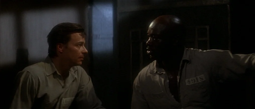 Assault on Precinct 13 (1976) movie still: medium — A dark, spartan space with muted grey walls and minimal interior detail. Two men in uniform…