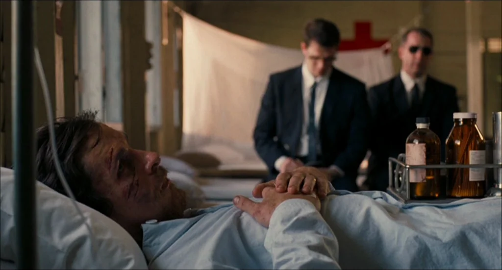 Rescue Dawn movie still: medium — A medical ward with white privacy screens and a red cross sign on the wall. A man lies in bed with injuries as two…