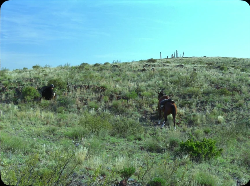 Jauja (2014) movie still: wide — A dry, grassy hillside landscape stretches out under a clear blue daylight sky. A lone man in a dark hat crouches…