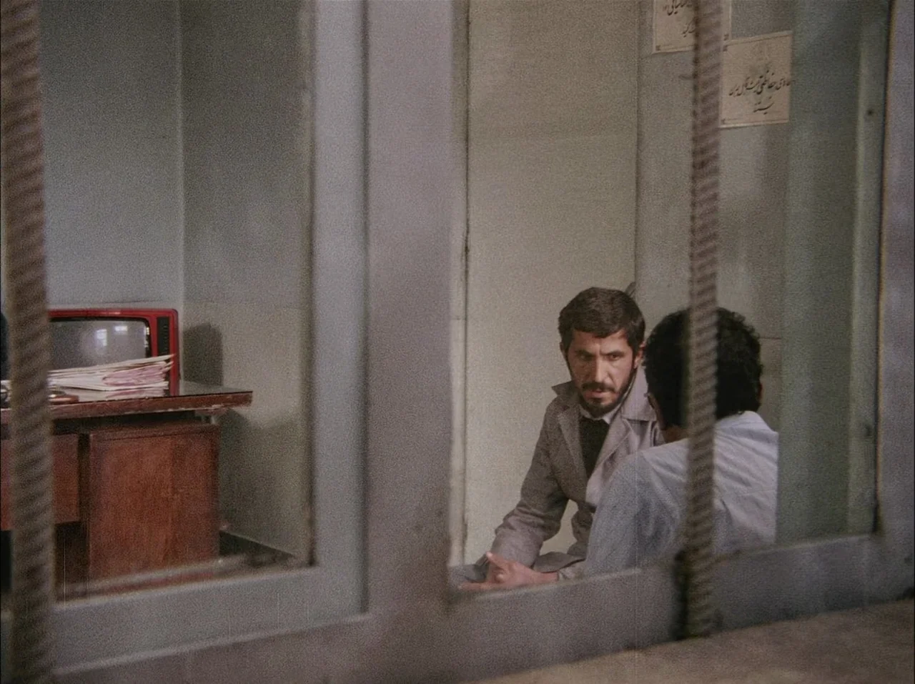 Close-Up (1990) movie still: medium — A stark visitation room with plain gray walls and metal frame partitions. Two men converse while sitting on…