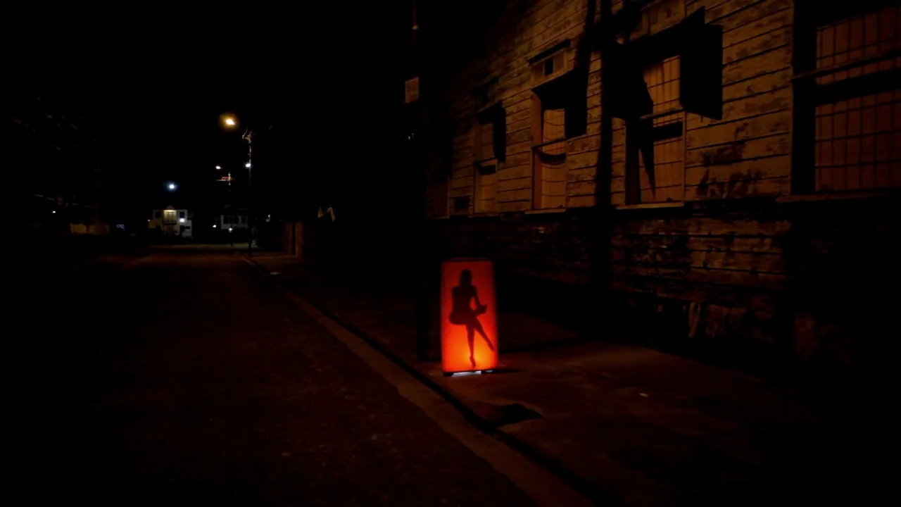 The ABCs of Death (2012) movie still: wide — A dark, empty street at night features a wooden building facade on the right. A single rectangular lamp…