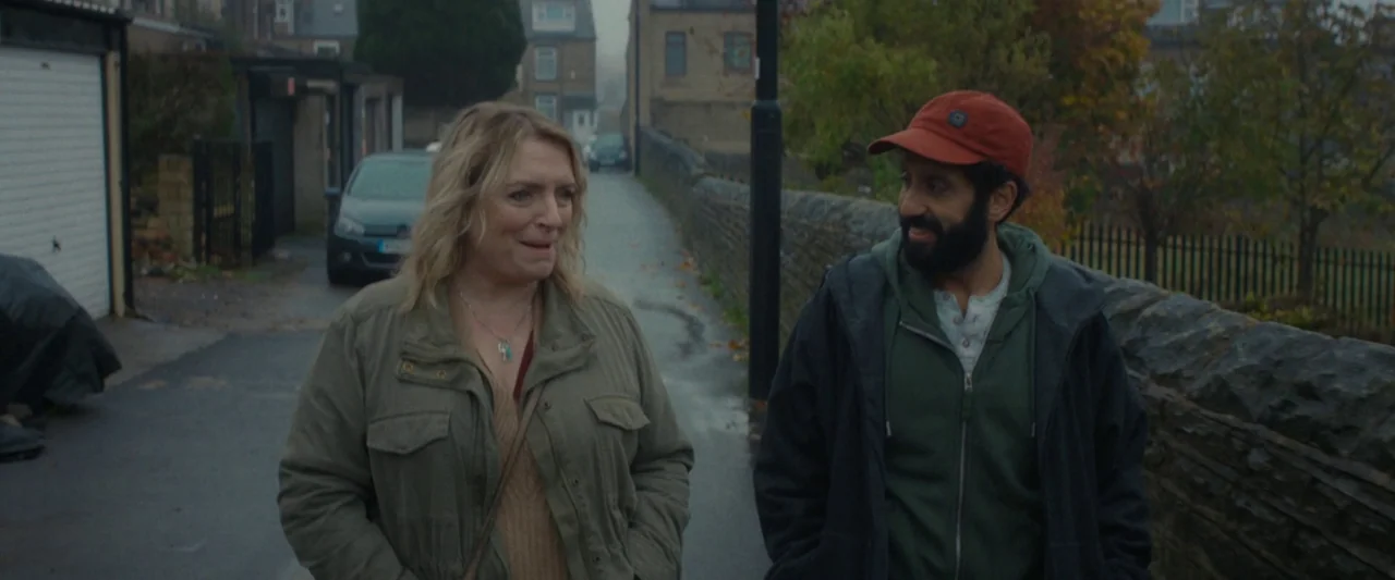 Ali & Ava (2021) movie still: medium — A quiet residential street lined with stone walls and garages on a cloudy day. A woman and a man walk…