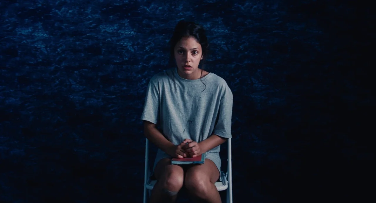 Ava (2017) movie still: medium — A woman sits on a simple white folding chair against a dark textured blue wall. She holds a small book on her lap…
