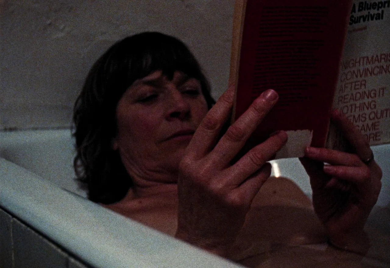 Enys Men (2022) movie still: closeup — A woman rests in a dimly lit white bathtub while reading a paperback book. Her eyes are closed as she holds…