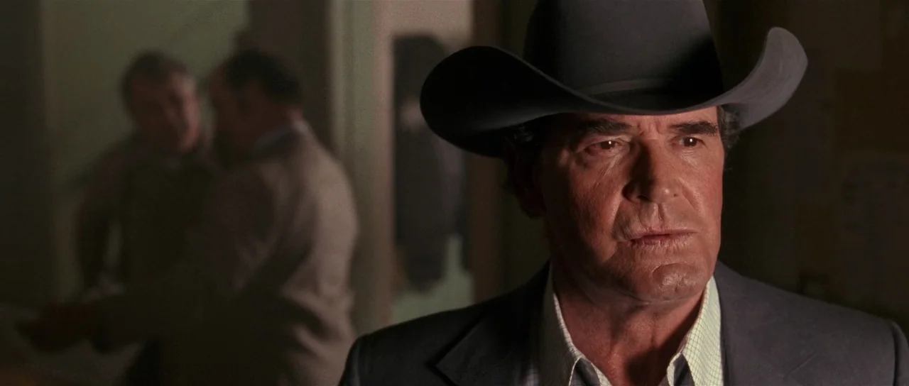 Fire In The Sky (1993) movie still: closeup — A man wearing a cowboy hat stands in the foreground of a dimly lit space. Two other blurred figures…
