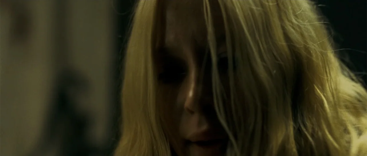 Blood Creek (2009) movie still: closeup — A dimly lit interior space with dark walls and heavy shadows. A blonde woman looks downward with her hair…