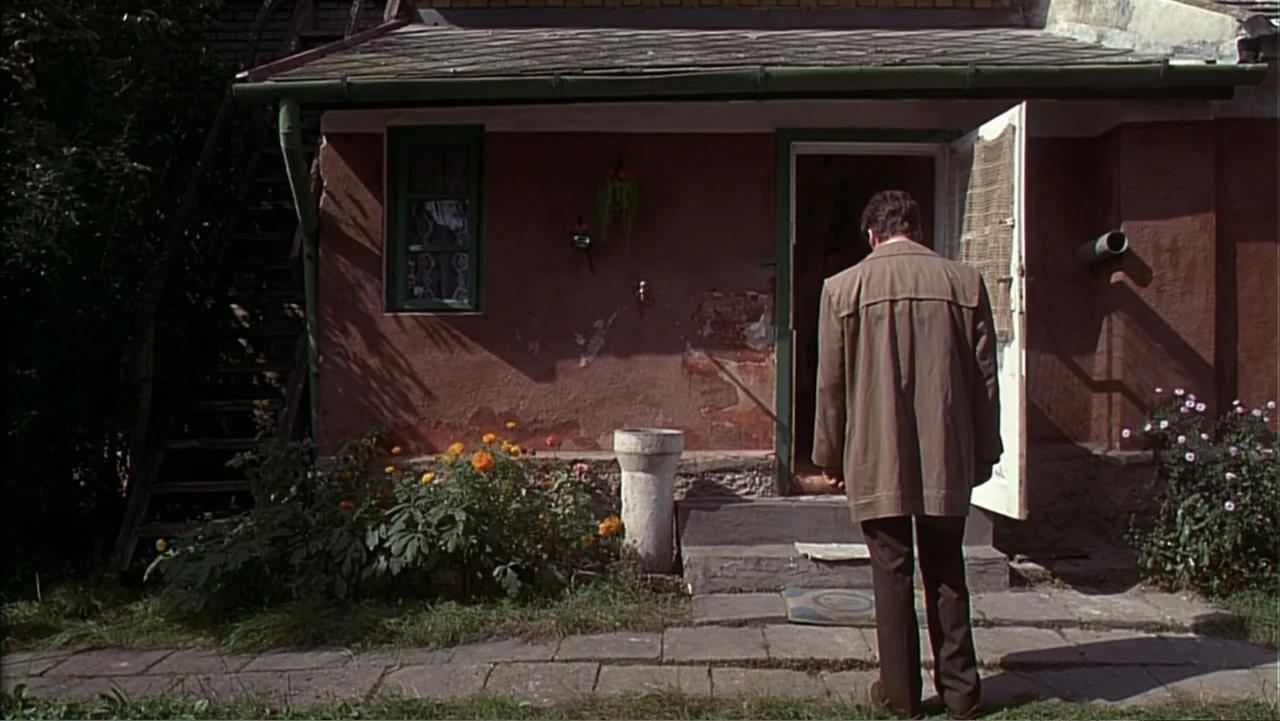 Citizen X (1995) movie still: fullbody — A weathered house exterior features red stucco, a concrete step, and marigold flower beds. A man in a brown…