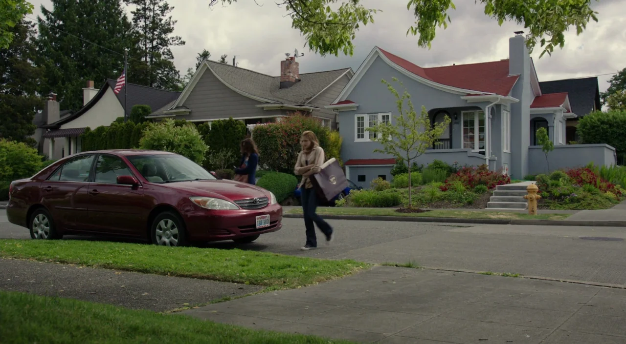 Laggies (2014) movie still: wide — A suburban street lined with detached houses and manicured lawns under an overcast sky. A woman walks past a red…