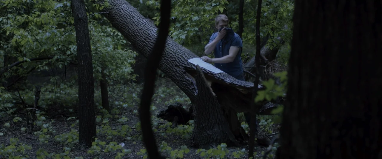 Radius (2017) movie still: medium — A dense patch of dark woods features a large fallen log. A man stands behind the log holding a document and…