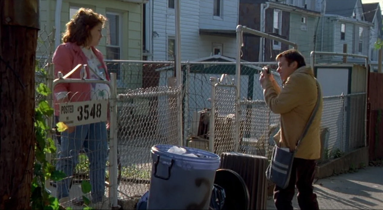 Pecker movie still: fullbody — A residential alleyway features rows of simple houses behind chain-link fences. A man uses a camera to take a picture…