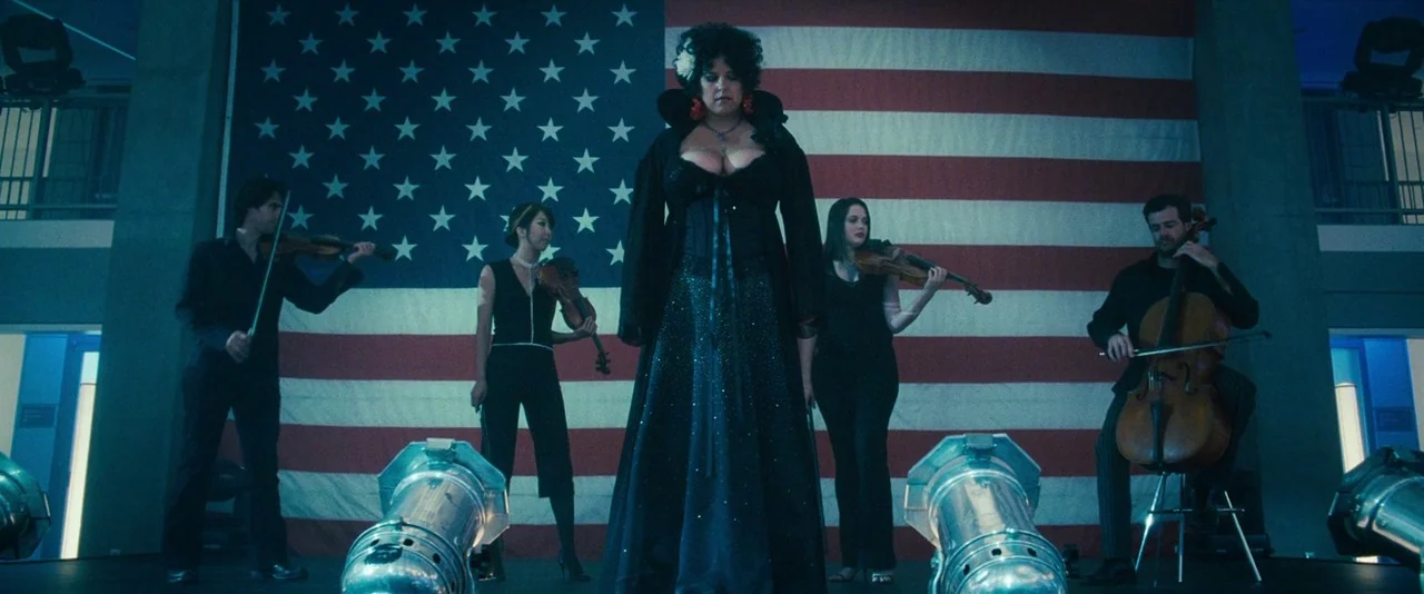 Southland Tales (2006) movie still: fullbody — A group of five musicians and a singer stand on a performance platform. A large American flag hangs…