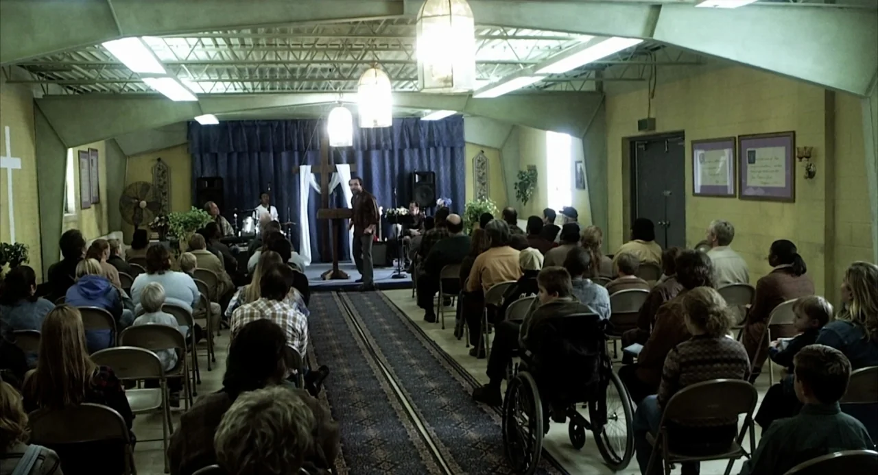 21 Grams (2003) movie still: wide — A multi-purpose room serving as a church with folding chairs and a small stage. A man stands behind a wooden…
