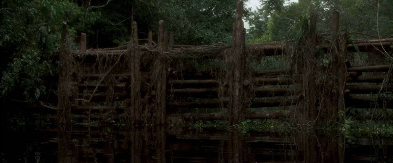 Anaconda (2025) movie still: establishing — A rustic wooden fence built from raw logs sits submerged in a calm river. Overgrown jungle foliage and…