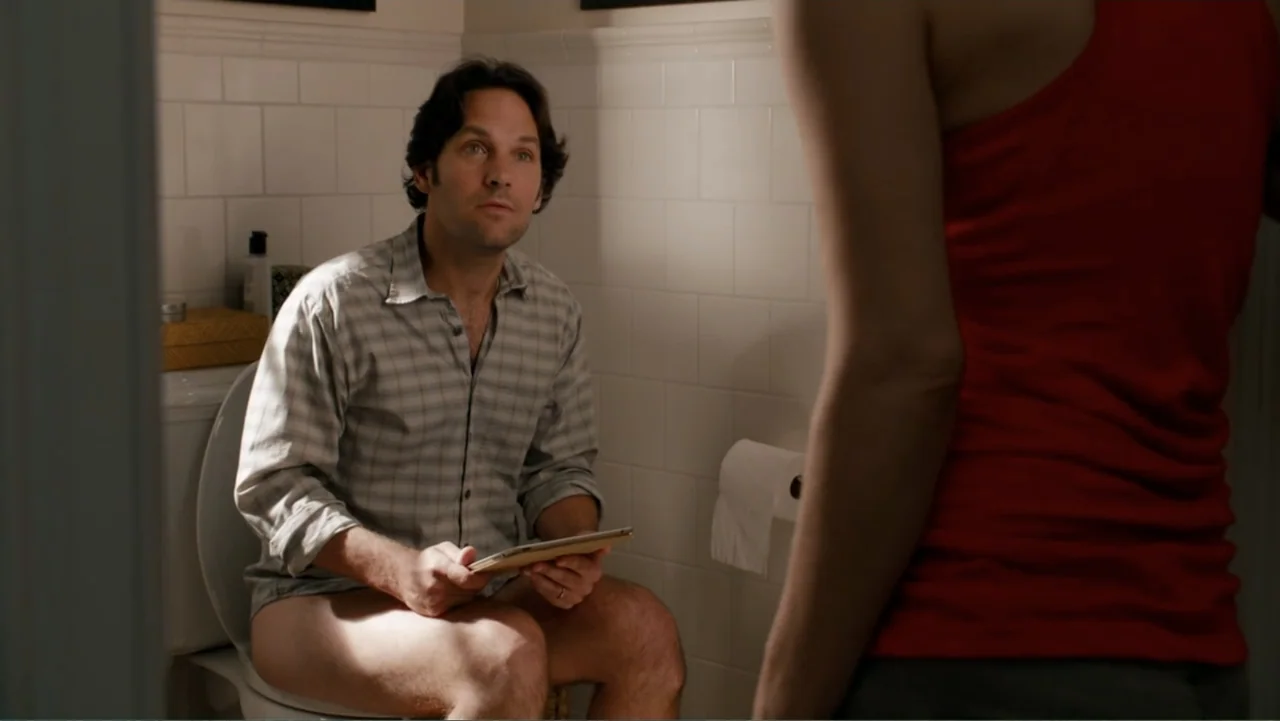 This Is 40 (2012) movie still: medium — A white tiled bathroom features a man seated on a toilet near the wall. He holds a digital tablet and looks…