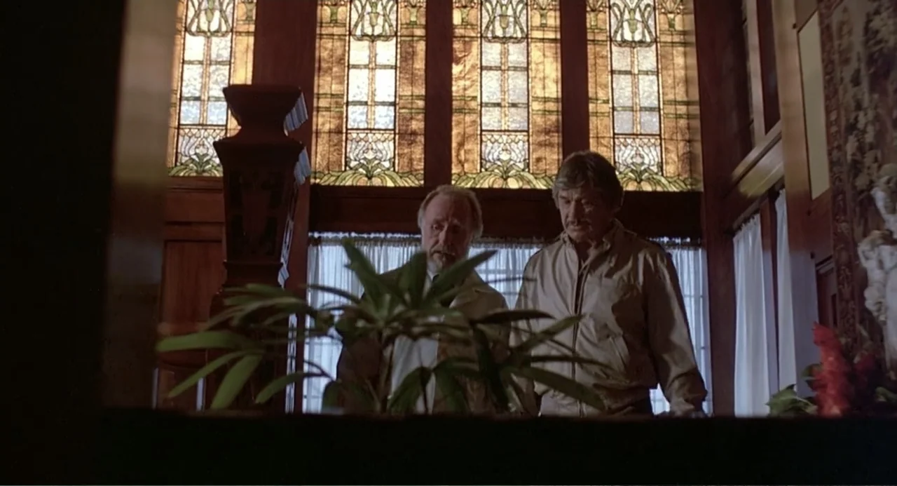 Death Wish II (1982) movie still: medium — An interior space features tall leaded glass windows with ornate patterns and wood trim. Two older men…