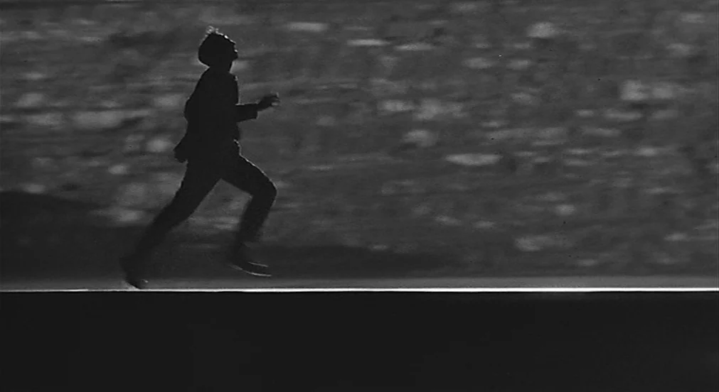 Kafka (1991) movie still: wide — A dark, empty road is seen at night under a bright horizon line. A lone man runs across the frame from left to…