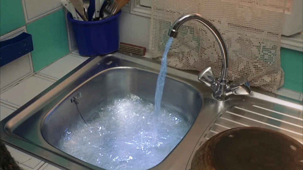 The Science Of Sleep (2006) movie still: closeup — A metal sink basin with white and turquoise tiles sits below a lace curtain. Water streams from a…
