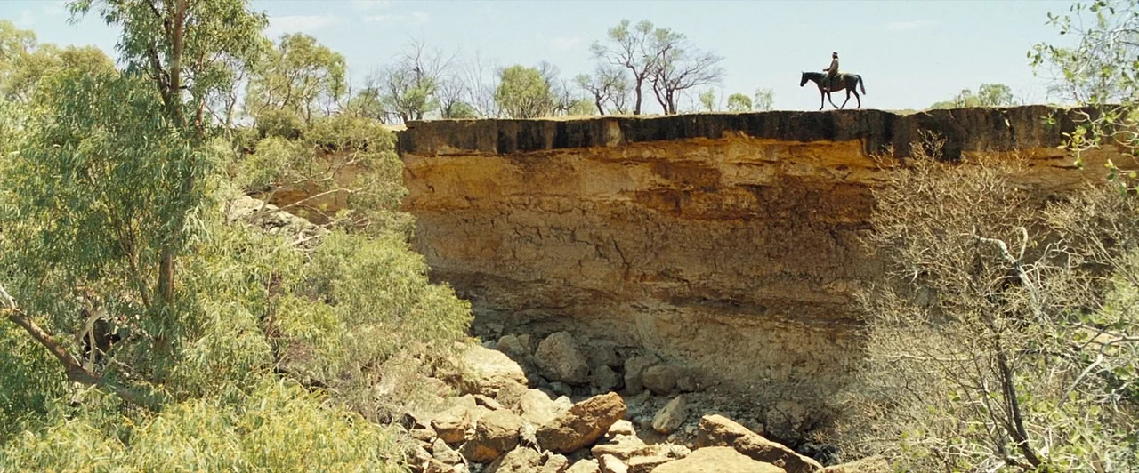 The Proposition (2005) movie still: wide — An arid, rocky Australian outback landscape features a tall, eroded sandstone cliff face. A single rider…